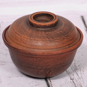 May include: A brown clay pot with a lid. The pot is round and has a smooth surface. The lid is slightly domed and has a small opening in the center.