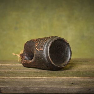Puede incluir: Una taza de cer&aacute;mica marr&oacute;n oscuro con un dise&ntilde;o decorativo en el lateral. La taza est&aacute; tumbada de lado sobre una superficie de madera.