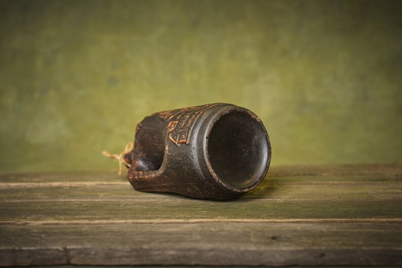 Puede incluir: Una taza de cer&aacute;mica marr&oacute;n oscuro con un dise&ntilde;o decorativo en el lateral. La taza est&aacute; tumbada de lado sobre una superficie de madera.