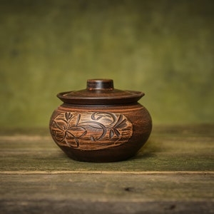 Op de afbeelding: Een handgemaakte, bruine keramische pot met deksel, op een houten oppervlak. De pot heeft een uitgesneden bloemenpatroon aan de zijkant. De achtergrond is een gedempt groen, wat een rustieke uitstraling creëert.