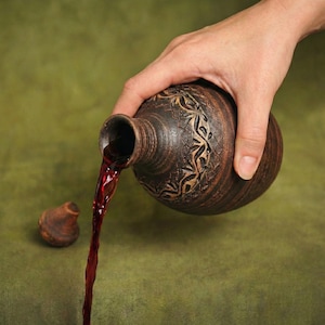 Può includere: Una bottiglia di ceramica marrone con un intricato disegno intagliato versa un flusso di liquido rosso. Un piccolo tappo rotondo è accanto. Lo sfondo è una superficie verde strutturata.