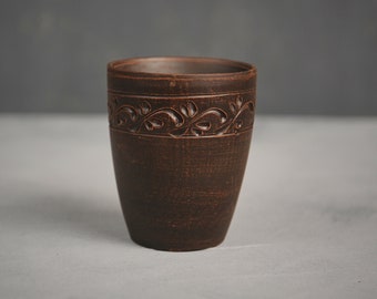 May include: A dark brown clay cup featuring a cylindrical shape with a slightly tapered base. The upper portion of the exterior is decorated with a carved, repeating vine and leaf pattern that encircles the entire circumference of the vessel.