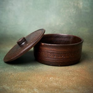 May include: A dark brown circular clay container with a matching lid. The exterior of the bowl and the top of the lid feature a carved, repeating floral vine pattern. The lid has a small, centered knob for lifting, and the bowl has a smooth, deep interior.