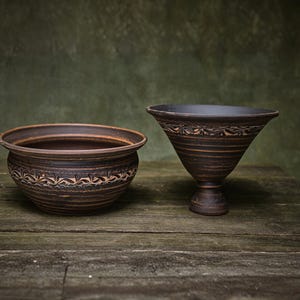 May include: A dark brown ceramic bowl with a rounded shape and a dark brown ceramic pedestal bowl with a wide, flared rim. Both pieces feature a horizontal band of carved leaf-like patterns around their midsections and a smooth, dark finish.