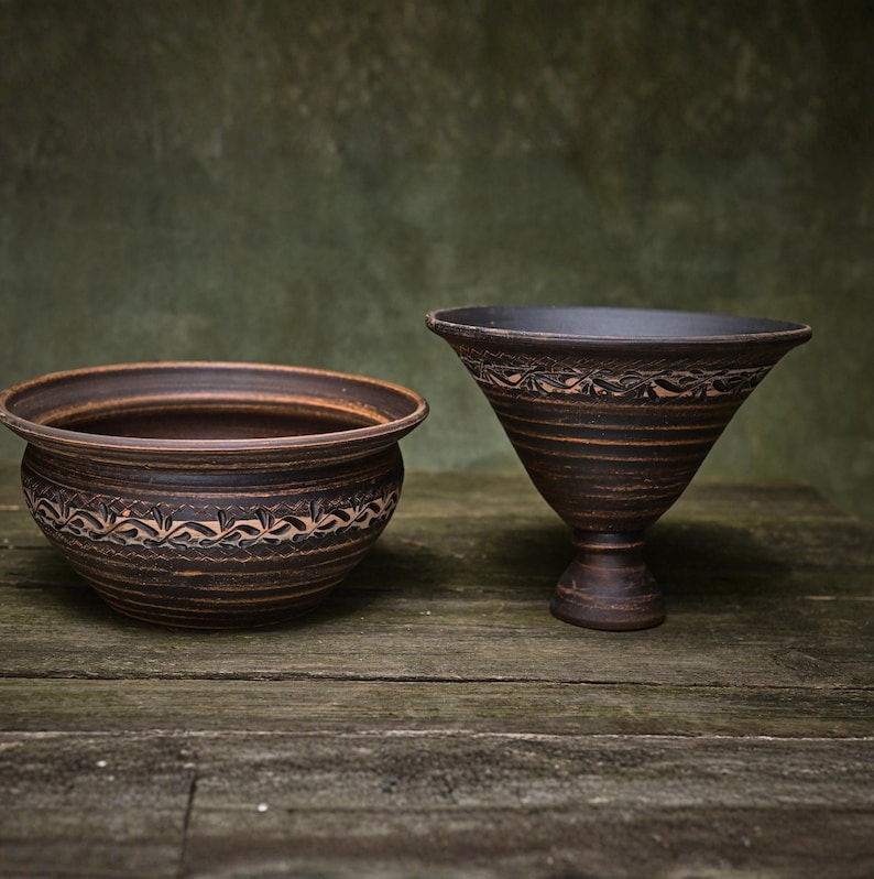 May include: A dark brown ceramic bowl with a rounded shape and a dark brown ceramic pedestal bowl with a wide, flared rim. Both pieces feature a horizontal band of carved leaf-like patterns around their midsections and a smooth, dark finish.