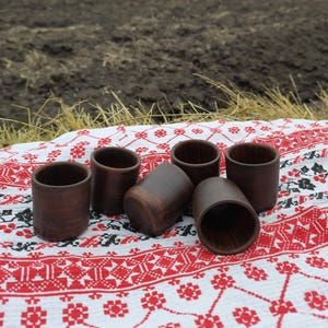Könnte beinhalten: Sechs kleine, braune Keramikbecher auf einem weißen Tuch mit rotem und schwarzem Blumenmuster. Die Becher sind zylindrisch mit einer strukturierten Oberfläche. Im Hintergrund ein Feld und Stroh.