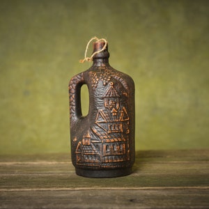 Puede incluir: Una botella de cer&aacute;mica marr&oacute;n oscuro con un dise&ntilde;o tallado de casas y vides. La botella tiene un asa y un tap&oacute;n de corcho. Est&aacute; sentada sobre una superficie de madera.