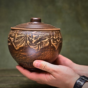 May include: A round, dark brown clay pot with a matching lid. The upper section of the pot features a textured, carved pattern in a lighter tan color. The surface of the pottery has a mottled, earthy finish with visible variations in the clay texture.