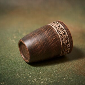 May include: A handcrafted, dark brown wooden cup with a decorative, carved band near the rim. The cup has a tapered shape and rests on a green and brown mottled surface. The carved band features a repeating floral design.