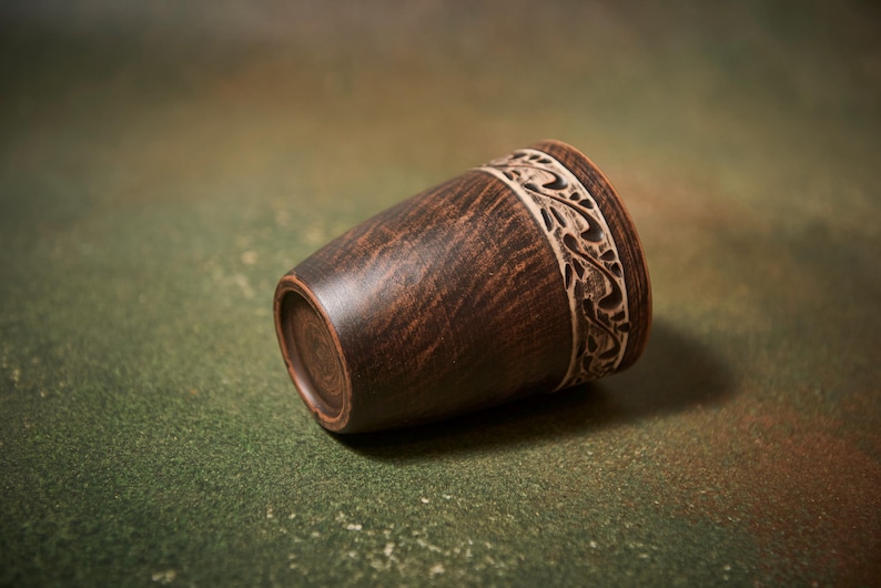 May include: A handcrafted, dark brown wooden cup with a decorative, carved band near the rim. The cup has a tapered shape and rests on a green and brown mottled surface. The carved band features a repeating floral design.