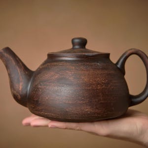 Peut inclure: Une théière en céramique marron avec un couvercle et une anse. La théière est tenue dans une main sur un fond marron. La théière a un corps arrondi et un bec verseur.