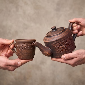 Könnte beinhalten: Eine Teekanne aus dunkelbraunem Ton und eine passende Tasse, beide mit aufwendigen, geprägten Schnörkelmustern verziert. Die Teekanne hat einen geschwungenen Ausguss, einen Deckel mit einem kleinen Knauf und einen Henkel, die Tasse einen Henkel.