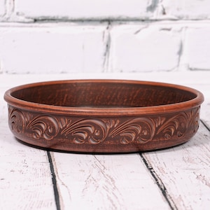 May include: A brown ceramic bowl with a decorative floral design around the rim. The bowl is sitting on a white wooden surface.