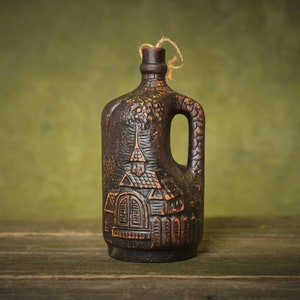Puede incluir: Una botella de cer&aacute;mica marr&oacute;n oscuro con asa y un dise&ntilde;o de una casa con un techo puntiagudo. La botella est&aacute; sentada sobre una superficie de madera.
