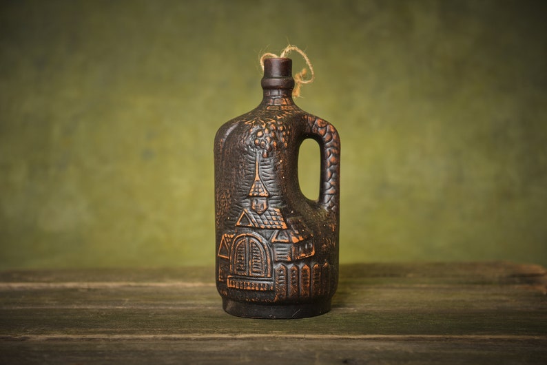 Puede incluir: Una botella de cer&aacute;mica marr&oacute;n oscuro con asa y un dise&ntilde;o de una casa con un techo puntiagudo. La botella est&aacute; sentada sobre una superficie de madera.