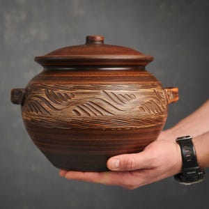May include: A round, dark brown clay pot with a matching lid and two side handles. The exterior features a wide, horizontal band of carved, repeating leaf-like patterns that wrap around the centre of the vessel, showcasing a textured finish.
