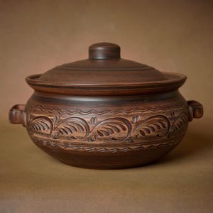 Könnte beinhalten: Ein brauner Keramiktopf mit Deckel, der mit einem dekorativen floralen Muster um die Mitte verziert ist. Der Topf hat zwei Griffe an den Seiten.