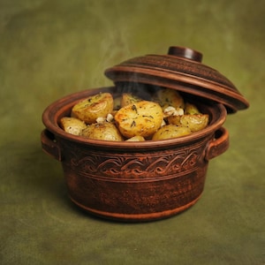 May include: A rustic, brown ceramic pot filled with roasted potatoes, with the lid slightly ajar. The potatoes are golden brown and seasoned, with steam rising from the pot. The pot has decorative carvings around the rim and two handles.