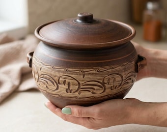 May include: A round brown clay pot with a matching lid and two small side handles. The body of the pot features a wide band of light brown horizontal texture decorated with a repeating carved swirl pattern around the center circumference.