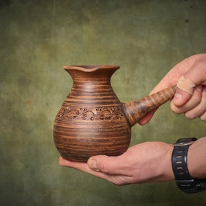 Könnte beinhalten: Eine handgefertigte, braune Keramik-Kaffeekanne mit langem Griff und dekorativem Blumendesign. Die Kanne wird in den Händen einer Person vor einem grünen Hintergrund gehalten. Die Kanne wird wahrscheinlich zum Brühen von Kaffee verwendet.