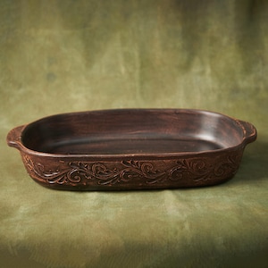 May include: A brown oval ceramic baking dish with a decorative floral design around the rim. The dish has two handles on either side.