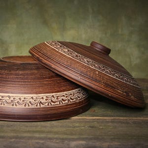 May include: A dark brown clay bowl and a matching lid, both featuring a horizontal band of carved leaf patterns. The bowl has a wide, circular base and the lid is domed with a small central knob, showcasing a natural, earthy texture.