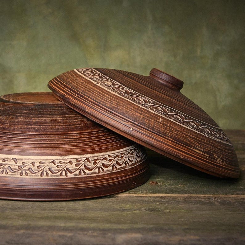 May include: A dark brown clay bowl and a matching lid, both featuring a horizontal band of carved leaf patterns. The bowl has a wide, circular base and the lid is domed with a small central knob, showcasing a natural, earthy texture.