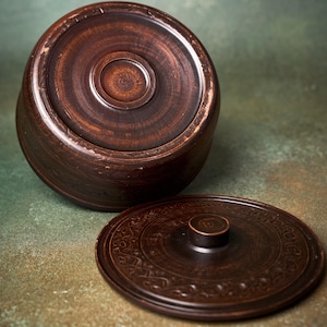 May include: A dark brown, round ceramic container with a matching lid. The container features a smooth, polished finish with visible circular textures. The lid has a central knob and an intricate, embossed floral pattern around the outer edge.