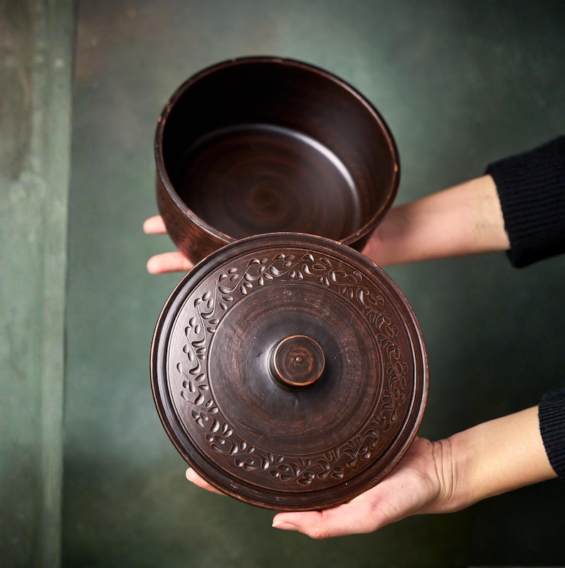 May include: A dark brown round clay pot with a matching lid. The lid features a circular embossed floral pattern and a central knob handle. The pot has a smooth, deep interior and a wide opening, showcasing the natural texture of the fired clay.