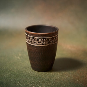 May include: A dark brown ceramic cup with a decorative band featuring a white floral design. The cup has a textured surface and a smooth, dark interior. The cup is likely used for drinking.