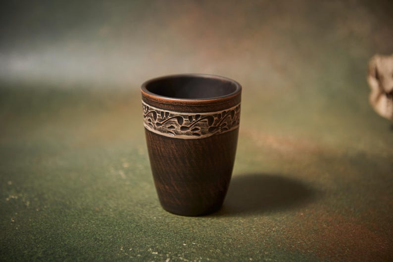 May include: A dark brown ceramic cup with a decorative band featuring a white floral design. The cup has a textured surface and a smooth, dark interior. The cup is likely used for drinking.