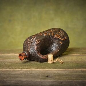 Puede incluir: Una botella de cer&aacute;mica marr&oacute;n con un tap&oacute;n de corcho, tumbada sobre una superficie de madera. La botella tiene una superficie texturizada con un dise&ntilde;o de uvas y hojas.