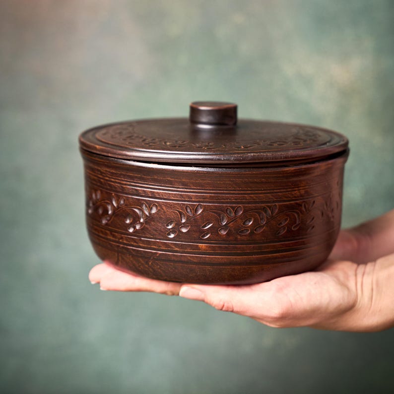May include: A round, dark brown ceramic bowl with a matching lid. The exterior of the bowl and the lid feature a carved, repeating vine and leaf pattern. The ceramic has a smooth, polished finish with a deep, rich, earthy color tone.