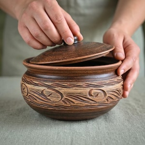 May include: A round, dark brown clay pot with a matching lid. The exterior features a textured, carved pattern of swirling lines and organic shapes encircling the body. The surface has a matte, earthy finish with variations in brown tones.