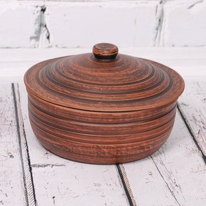 Può includere: Un vaso di ceramica marrone con un coperchio. Il vaso è rotondo e decorato con una serie di cerchi concentrici.