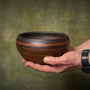 May include: A round, deep bowl crafted from dark brown clay with a smooth, matte finish. The exterior features subtle, horizontal ridged textures and a gradient of deep espresso tones blending into warmer, earthy terracotta hues along the sides.