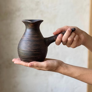 May include: A dark brown ceramic coffee pot with a rounded body, a flared rim, and a long side handle. The surface features horizontal textured lines and a small diamond-shaped mark near the base, showcasing a rustic, unglazed appearance.