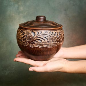Può includere: Un vaso in argilla marrone scuro di forma rotonda con coperchio abbinato. L'esterno presenta un motivo a foglie intagliato a mano che circonda la parte superiore, mentre il coperchio mostra creste circolari concentriche.