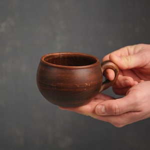 May include: A brown ceramic mug with a rounded body and a curved handle. The mug is held in two hands against a grey background. The mug appears to be made of clay and has a rustic, natural appearance. The mug is approximately 10 cm in diameter.