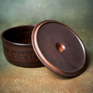 May include: A dark brown ceramic bowl with a matching circular lid. The bowl features a decorative embossed band around its exterior. The lid is smooth with a central circular indentation. The ceramic has a deep, earthy tone with subtle variations in color throughout the surface.