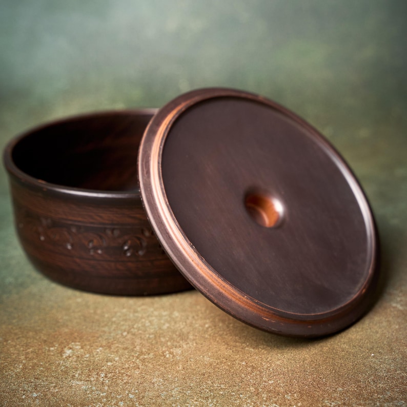 May include: A dark brown ceramic bowl with a matching circular lid. The bowl features a decorative embossed band around its exterior. The lid is smooth with a central circular indentation. The ceramic has a deep, earthy tone with subtle variations in color throughout the surface.