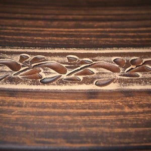 May include: A dark brown ceramic surface featuring a horizontal band with a carved leaf pattern. The carved design reveals a lighter beige color beneath the dark brown glaze, creating a contrast between the textured floral motif and the smooth finish.