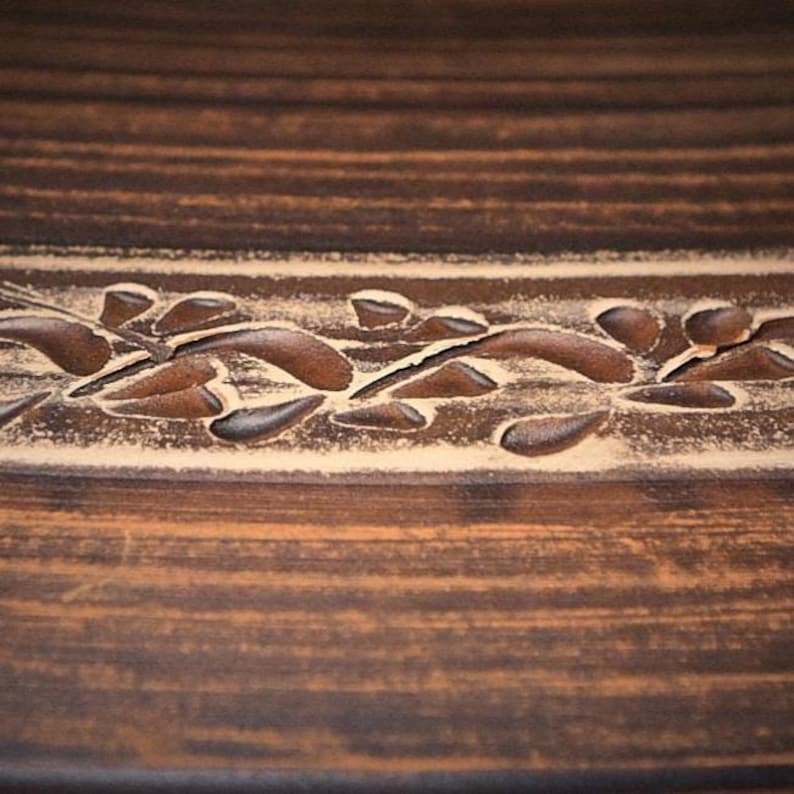 May include: A dark brown ceramic surface featuring a horizontal band with a carved leaf pattern. The carved design reveals a lighter beige color beneath the dark brown glaze, creating a contrast between the textured floral motif and the smooth finish.