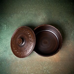 May include: A round, dark brown clay pot with a matching lid. The lid features a central knob and a decorative embossed floral pattern around the perimeter. The pot has a smooth, deep interior and a wide, open circular rim.