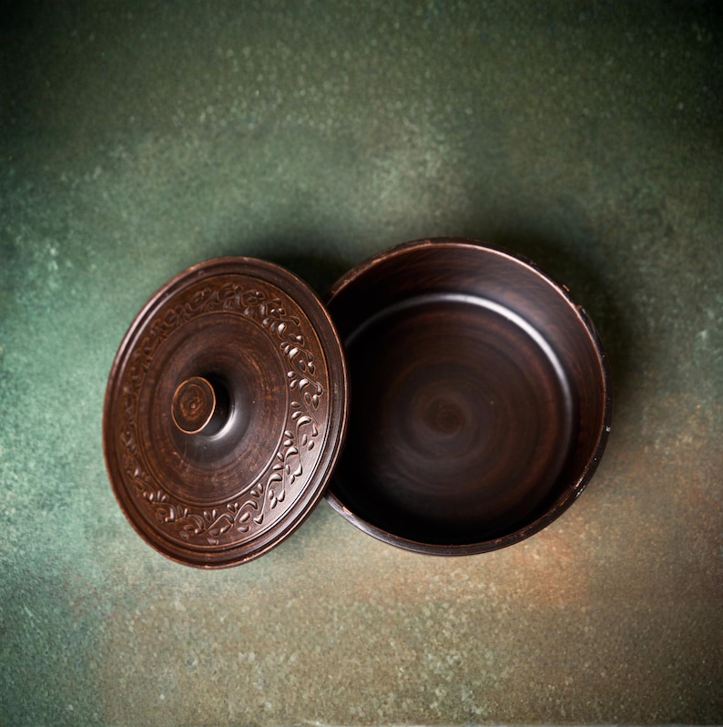 May include: A round, dark brown clay pot with a matching lid. The lid features a central knob and a decorative embossed floral pattern around the perimeter. The pot has a smooth, deep interior and a wide, open circular rim.