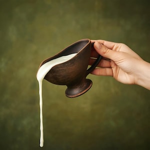 May include: A brown ceramic gravy boat with a black handle, filled with white liquid, is being poured. The gravy boat has a wide, open top and a pedestal base. The liquid is flowing out of the boat in a thick stream against a green background.