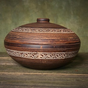 Peut inclure: Un bol en céramique marron avec un couvercle. Il présente des motifs floraux crème et des lignes horizontales. Le couvercle est orné d'un petit bouton. Le bol est posé sur une surface en bois. Diamètre: environ 25 cm.