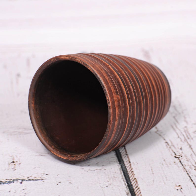 May include: A cylindrical brown clay cup featuring a series of horizontal, raised ridges encircling the exterior. The interior of the cup is smooth and dark brown, while the outer surface displays a textured, ribbed pattern in earthy tones.