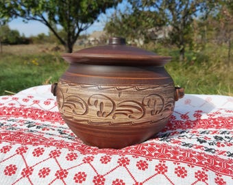 May include: A brown ceramic pot with a lid, featuring carved decorative patterns. The pot has two small handles and sits on a white and red patterned cloth, suggesting a rustic or farmhouse aesthetic.