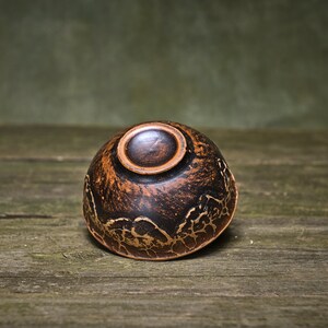 May include: A small, dark brown ceramic bowl with a gold-colored design. The bowl is sitting on a wooden surface.